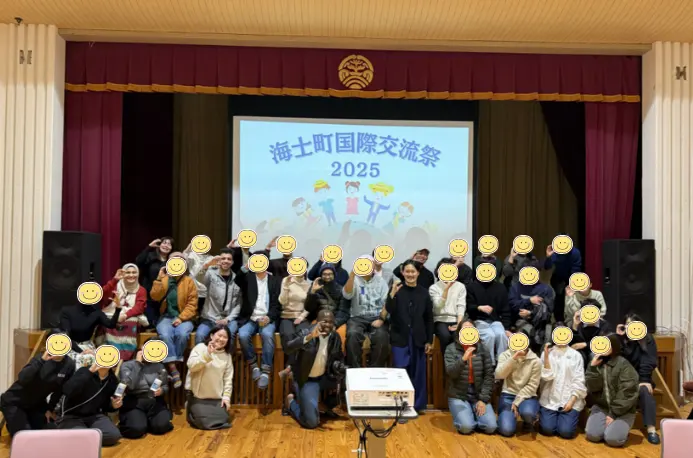 A large group of festival participants on a stage under a banner for the Ama Town International Exchange Festival 2025; faces are covered with smiley icons.