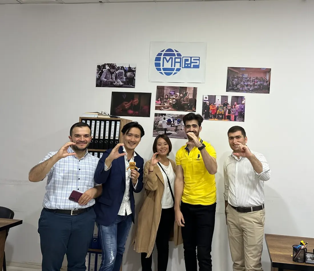 Five people standing together in an office in front of a wall of photographs, each making a C shape with one hand.